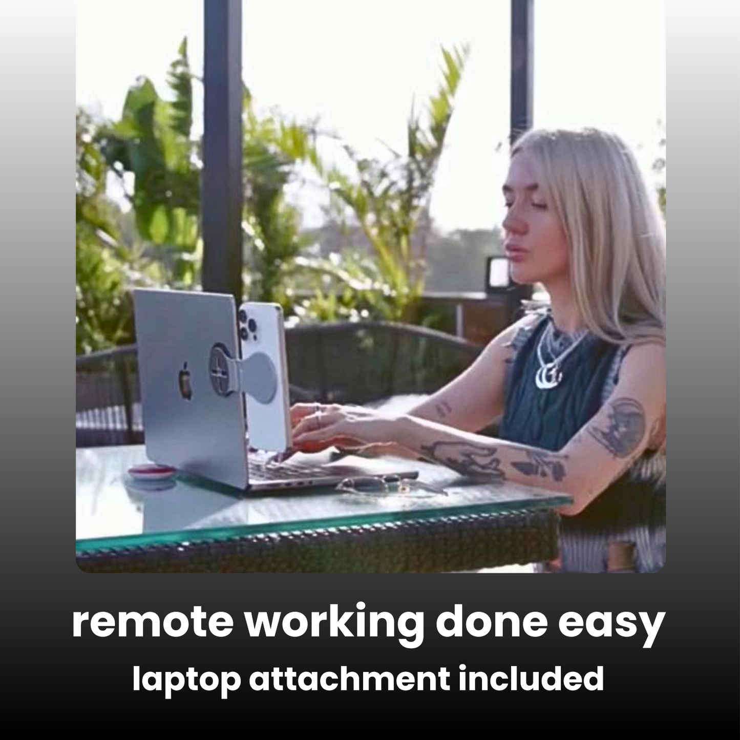A woman with blonde hair and tattoos sits at a glass table outdoors, using a silver laptop with a circular magnetic attachment. The image includes text at the bottom reading 'remote working done easy laptop attachment included'.